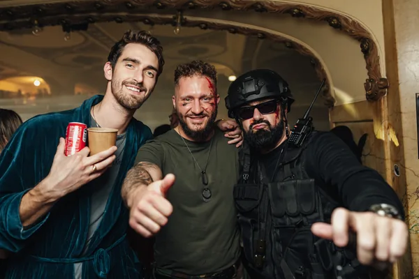 Tři hosté v kostýmech pózují a gestikulují směrem k fotoaparátu